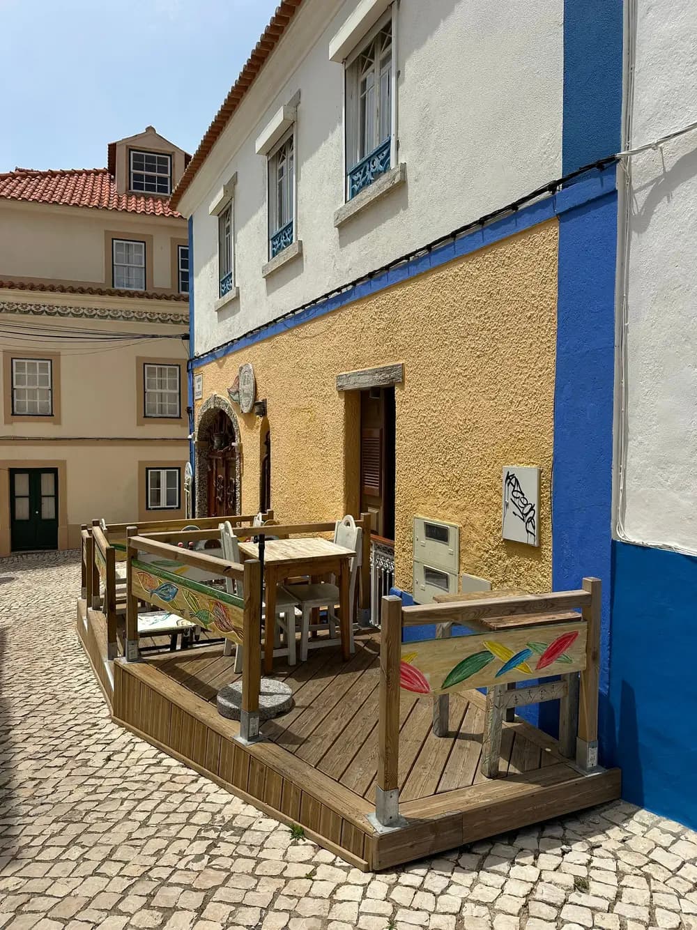 Fachada branca com barra azul e mesas de madeira à porta